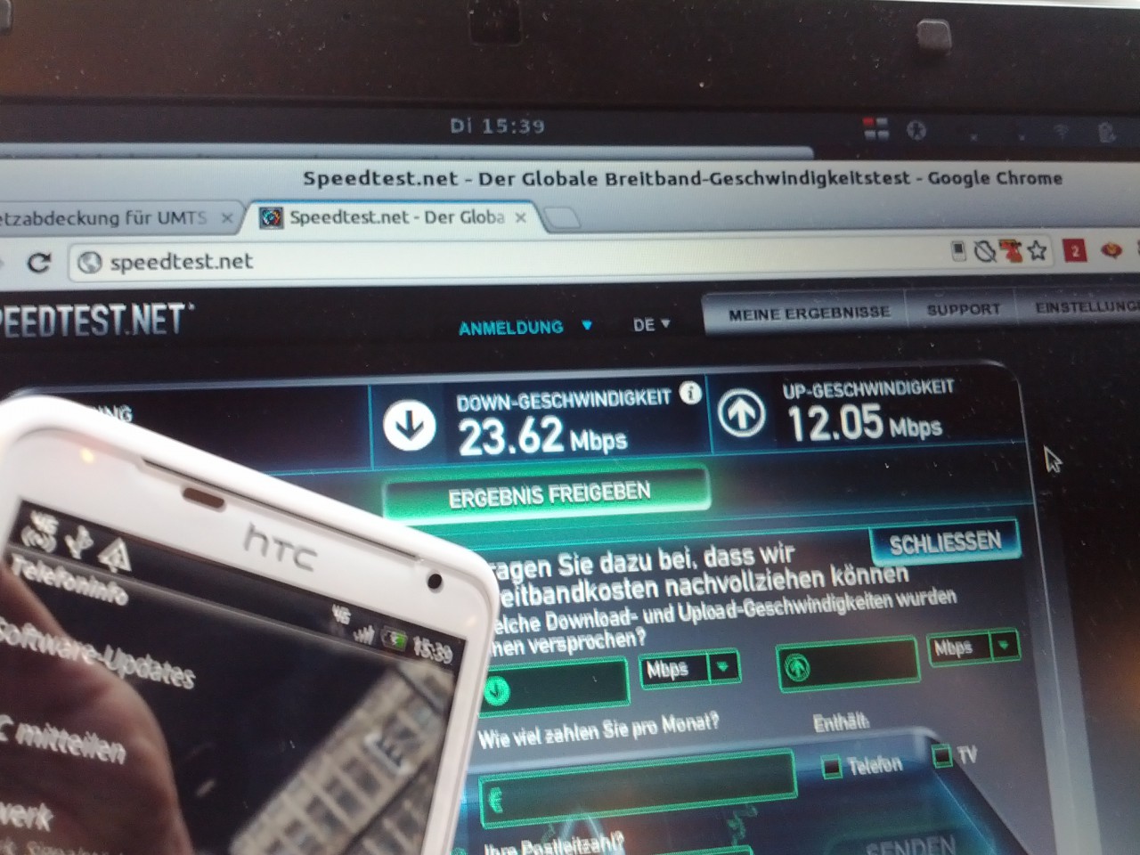 Ein weißes HTC-Handy vor einem Notebook, auf dem Speedtest.net 23,62 MBit/s im Download und 12,05 MBit/s im Upload anzeigt
