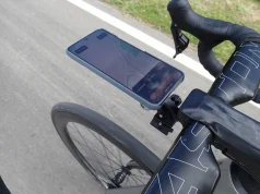 Nahaufnahme der Rennradlenker mit einem Smartphone, das in einer Halterung am Vorbau befestigt ist. Auf dem Handybildschirm ist eine Navigations-App mit einer geplanten Route zu sehen, die den aktuellen Weg des Radfahrers anzeigt. Die Lenker sind mit schwarzem Lenkerband umwickelt, besitzen integrierte Bremshebel und ein aerodynamisches Design. Im Hintergrund erkennt man die Asphaltstraße sowie einen Grünstreifen am Fahrbahnrand, was auf eine Fahrradtour im Freien hinweist. Keywords: Rennrad Handyhalterung, Fahrrad GPS Navigation, Smartphone Lenkerhalter, Rennrad Zubehör, Fahrrad Navigation.