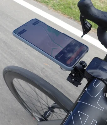 Nahaufnahme der Rennradlenker mit einem Smartphone, das in einer Halterung am Vorbau befestigt ist. Auf dem Handybildschirm ist eine Navigations-App mit einer geplanten Route zu sehen, die den aktuellen Weg des Radfahrers anzeigt. Die Lenker sind mit schwarzem Lenkerband umwickelt, besitzen integrierte Bremshebel und ein aerodynamisches Design. Im Hintergrund erkennt man die Asphaltstraße sowie einen Grünstreifen am Fahrbahnrand, was auf eine Fahrradtour im Freien hinweist. Keywords: Rennrad Handyhalterung, Fahrrad GPS Navigation, Smartphone Lenkerhalter, Rennrad Zubehör, Fahrrad Navigation.