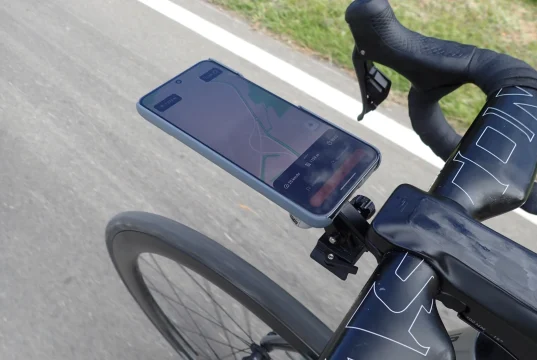 Nahaufnahme der Rennradlenker mit einem Smartphone, das in einer Halterung am Vorbau befestigt ist. Auf dem Handybildschirm ist eine Navigations-App mit einer geplanten Route zu sehen, die den aktuellen Weg des Radfahrers anzeigt. Die Lenker sind mit schwarzem Lenkerband umwickelt, besitzen integrierte Bremshebel und ein aerodynamisches Design. Im Hintergrund erkennt man die Asphaltstraße sowie einen Grünstreifen am Fahrbahnrand, was auf eine Fahrradtour im Freien hinweist. Keywords: Rennrad Handyhalterung, Fahrrad GPS Navigation, Smartphone Lenkerhalter, Rennrad Zubehör, Fahrrad Navigation.