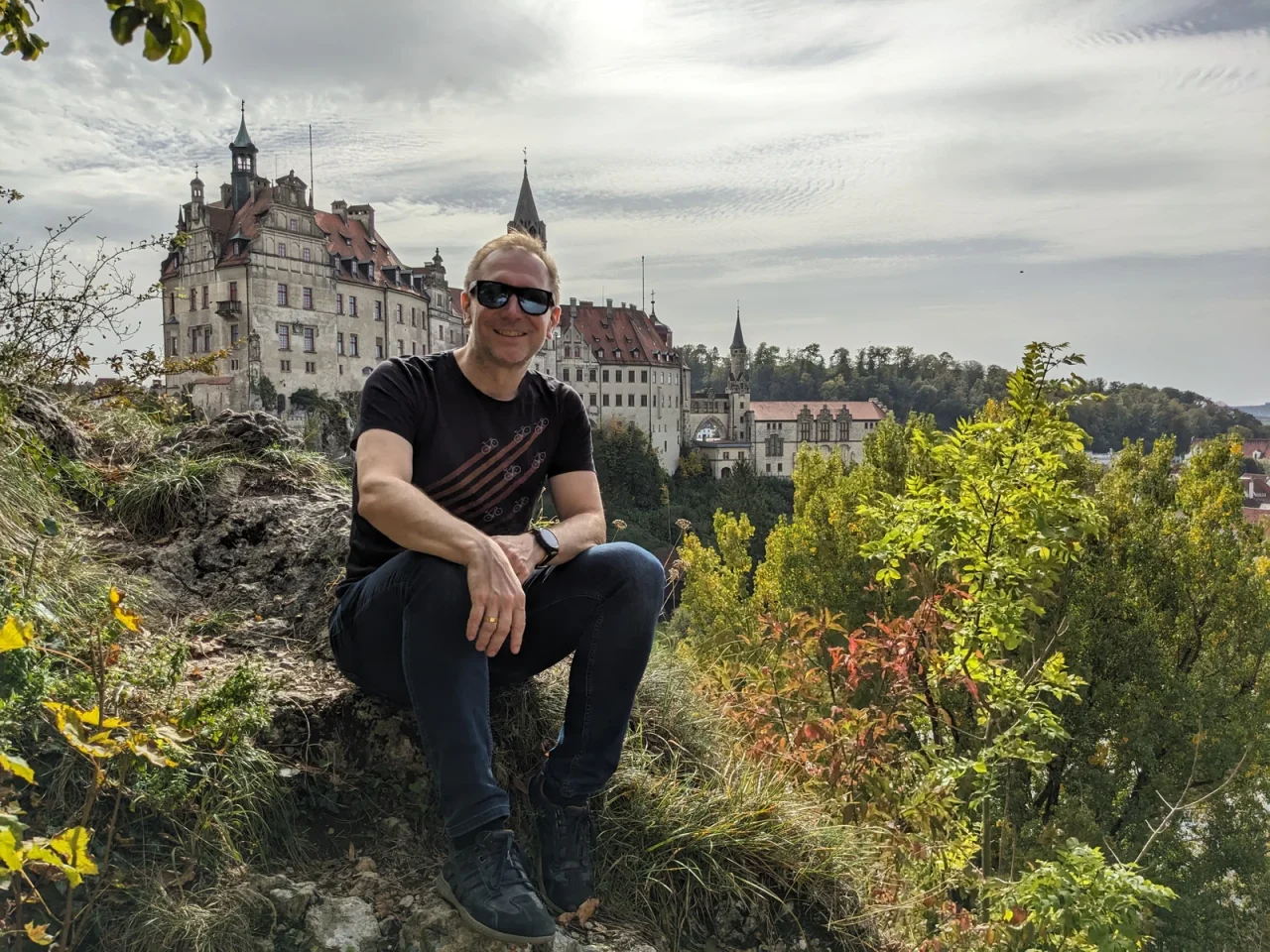 Christoph mit Sonnenbrille sitzt auf einem Felsen, im Hintergrund eine große Schlossanlage zwischen bewaldeten Hügeln.
