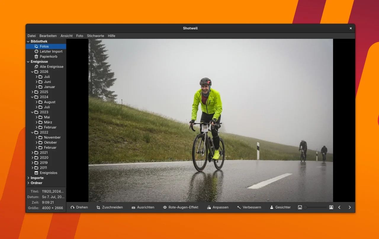 Das Programmfenster der Fotoverwaltungssoftware Shotwell präsentiert eine Aufnahme eines lächelnden Rennradfahrers in leuchtend gelber Jacke auf einer nassen Asphaltstraße. Im Hintergrund des nebligen Landschaftsmotivs sind weitere Radfahrer sowie ein grasbewachsener Hang zu sehen. Auf der linken Seite der Benutzeroberfläche befindet sich eine Ordner-Bibliothek, während am unteren Rand Werkzeuge zur Bildbearbeitung angeordnet sind.